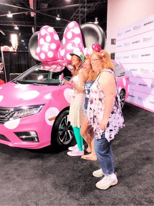 3 girls at the D23 Expo in front of a pink and white car with minnie ears on top of it
