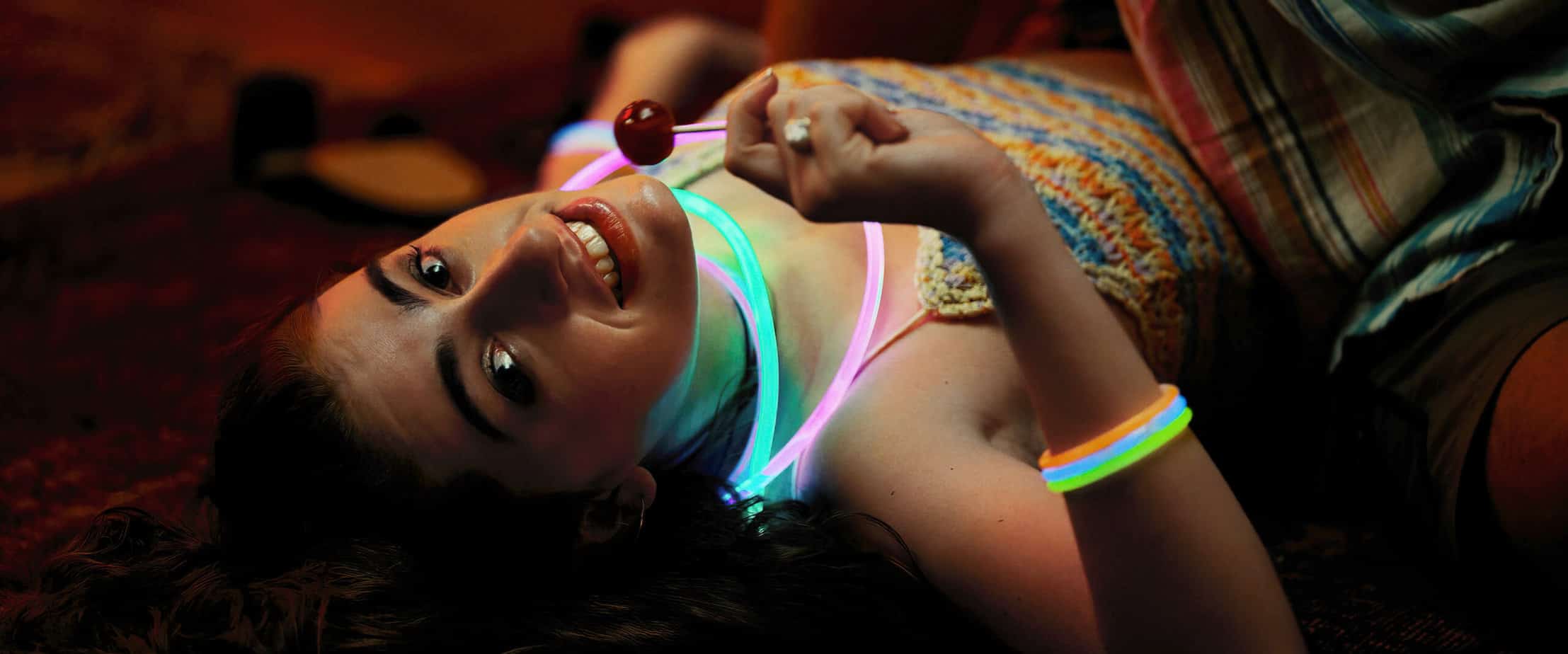Age Rating for Bodies Bodies Bodies parents guide. Girl laying on her back looking at the camera with glow necklaces around her neck. She is holding a blow pop lollipop.