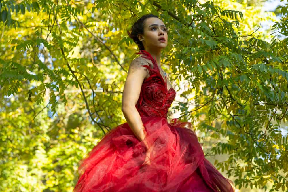 The Invitation pee breaks. Woman outside with trees in the background. She is wearing a gorgeous red ballgown and looks scared.