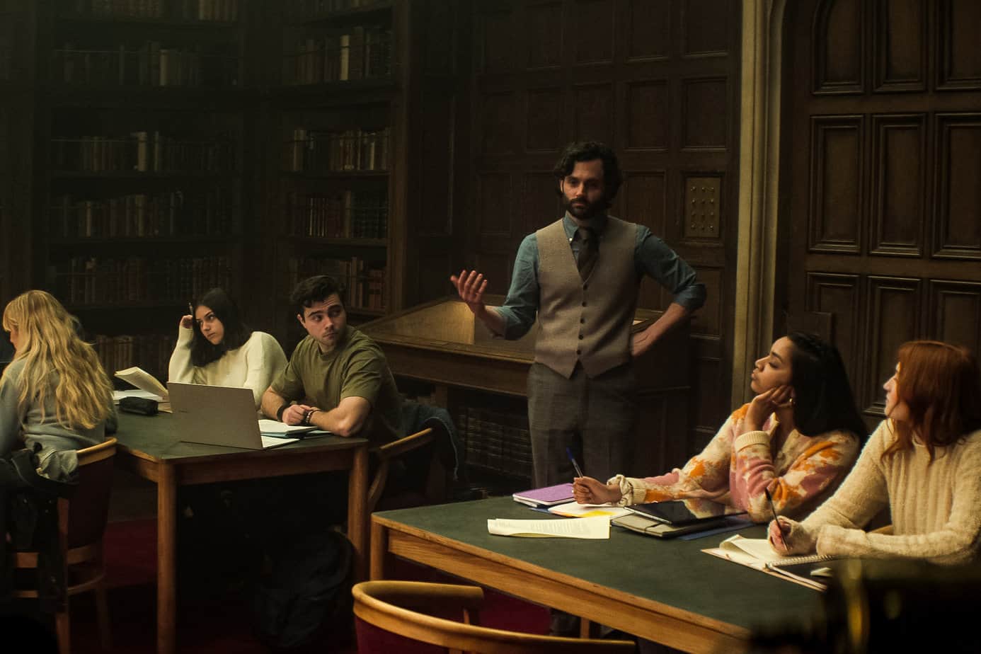 Age rating of You season 4 netflix parents guide. Professor in a library with students teaching a class.